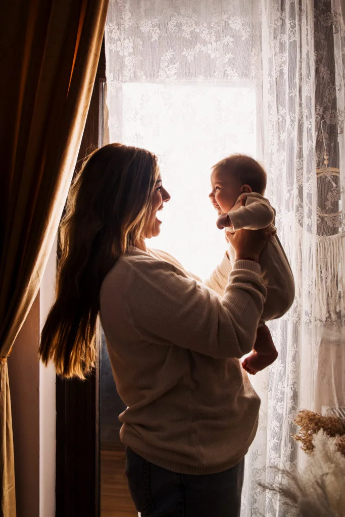 Mamă și bebeluș fericiți la fereastră, ședință foto de familie călduroasă București