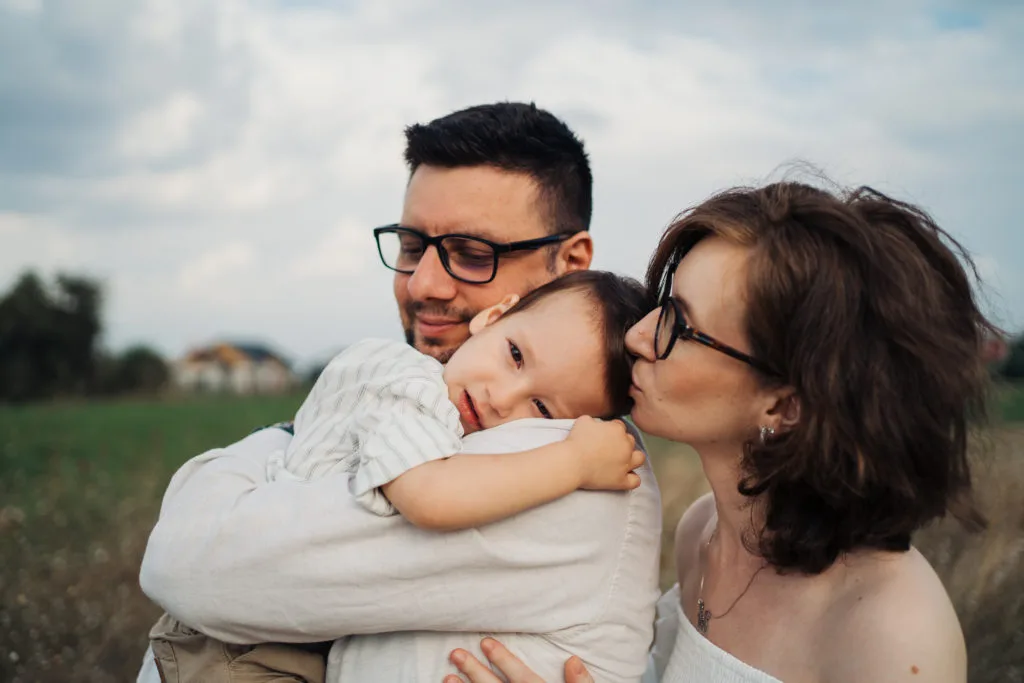 Ședință foto familie în exterior, părinți cu ochelari îmbrățișând copil mic pe câmp