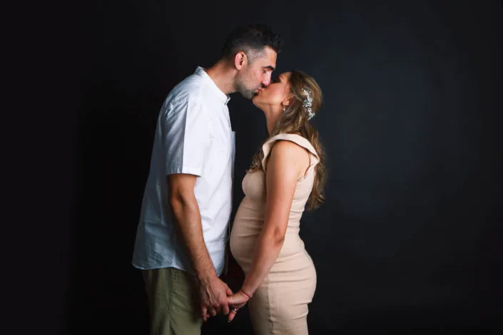 Cuplu în ședință foto maternitate, sărut romantic în studio cu fundal întunecat