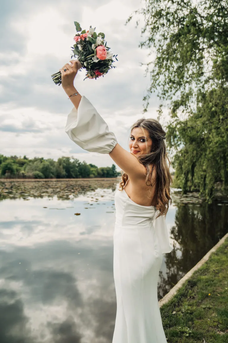 Mireasă veselă cu buchet ridicat lângă lac, fotografie de nuntă în aer liber