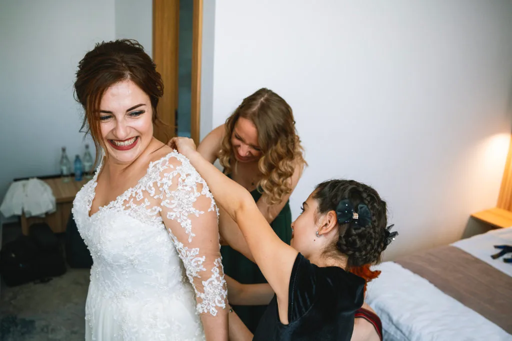 Mireasă râzând în timp ce domnișoarele de onoare o ajută cu rochia, fotografie nuntă