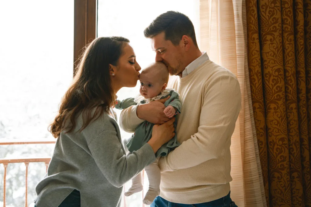 Ședință foto familie cu bebeluș, părinți sărutând copilul în lumină caldă naturală