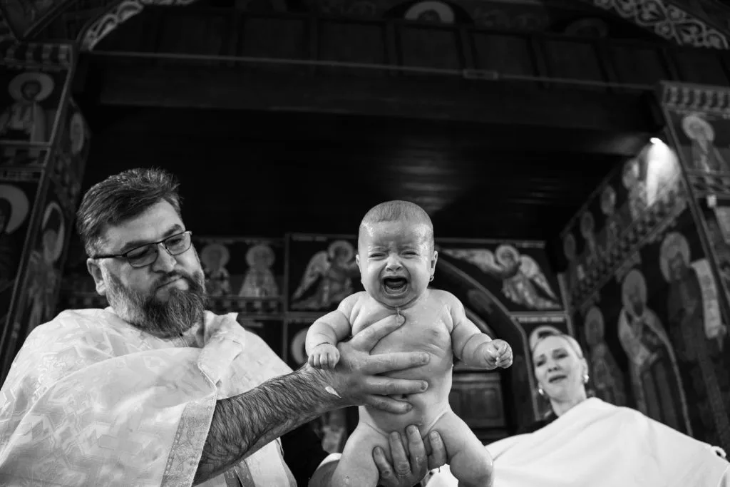 Bebeluș plângând după botez ortodox ținut de preot în biserică decorată cu icoane