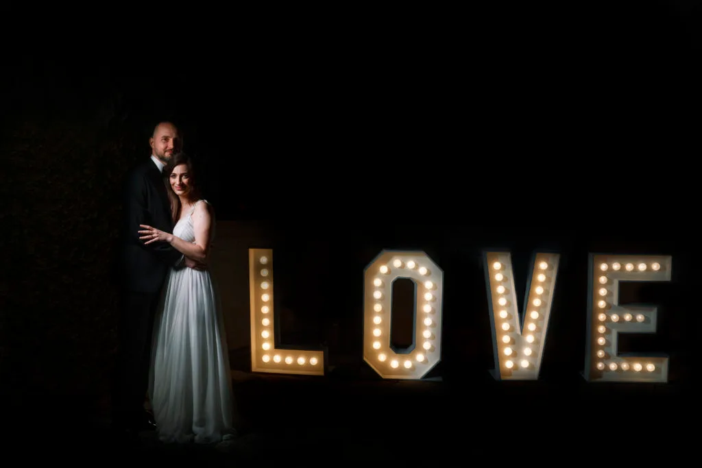 Miri îmbrățișați lângă litere luminoase LOVE, fotografie romantică de nuntă București