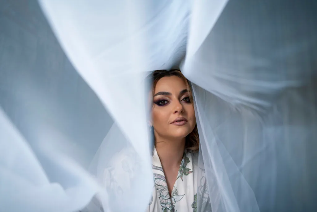 Ședință foto portret femeie privind printre voaluri albe translucide atmosferă artistică