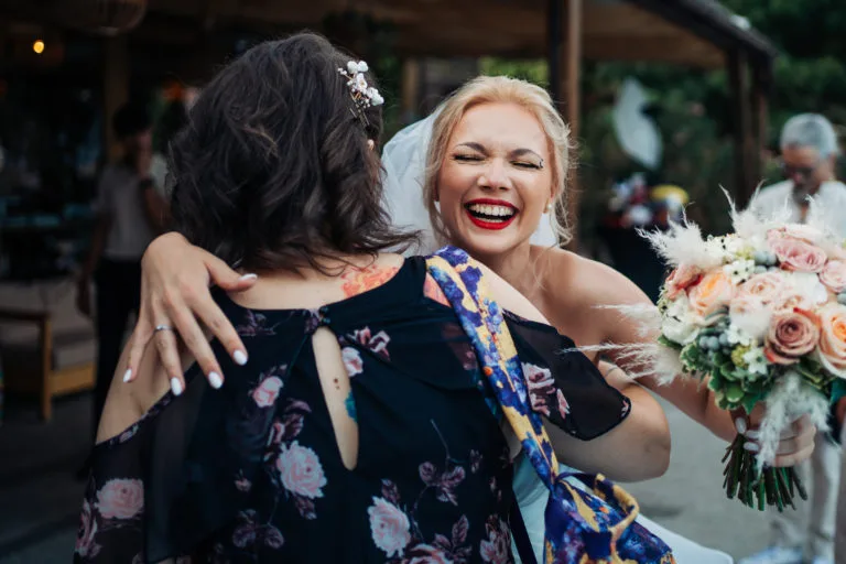 Mireasă râzând și îmbrățișând o invitată cu buchet de flori, fotografie de nuntă